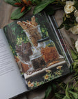 Open book with a decorative cover featuring floral designs, surrounded by green leaves and white flowers on a textured surface.