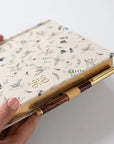 Hand holding a floral notebook with a pen holder on a white background