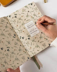 Person holding a floral notebook with a pen, on a white surface with a yellow pouch and pens.