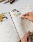 Person using a planner with floral designs and a hand holding a small object.