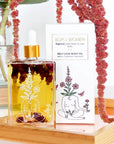 Bottle of self-love body oil with a dropper on a wooden stand, surrounded by pink flowers.