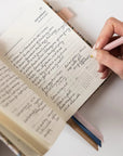 Person writing in a notebook with a pink pen on a white surface