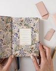 Floral-patterned notebook held by hands with a pen and pink case on a light background