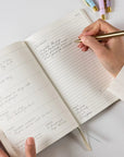 Person writing in a notebook with a gold pen on a light surface.