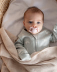 Baby in a light gray onesie with a pacifier, lying on a beige blanket in a woven basket.