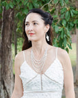 Woman in a white dress with pearl necklace and earrings standing in a natural setting.