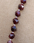 Faceted dark red gemstone beads strung on a white cord, displayed on a textured beige fabric background.