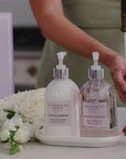 Two bottles of Peppermint Grove hand and body wash on a tray with flowers and a blurred background.