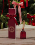Red diffuser bottle with matching packaging on a wooden surface with Christmas decorations in the background