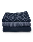 Three folded knit blankets in shades of navy and slate blue, stacked on a white background. The top blanket features a geometric pattern, and the middle and bottom are solid textured knits.