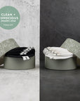 Two round metal gift boxes with patterned lids, each containing a bundle of elastic hair ties. The left box shows black hair ties with silver metal ends, and the right box shows white hair ties with black ends.