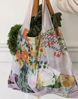 Reusable shopping bag with floral and fruit design filled with groceries, held by a person.