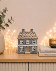 A small gray ceramic house figurine sits on a light wood cabinet, styled with a beige ribbed vase holding eucalyptus leaves on the left and a stack of books with a lit candle on the right against a warm beige wall with string lights.