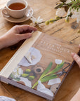 Person holding a book titled 'After Hours' on a wooden table with a cup of tea and flowers.