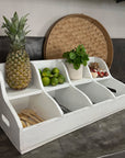 White wooden fruit basket with compartments containing a pineapple, limes, and other items on a kitchen counter.
