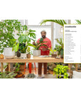 Person in a room filled with plants, holding a plant, with a table and book visible.