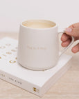 White mug with 'Hug in a Mug' text held over an open book on a light surface.