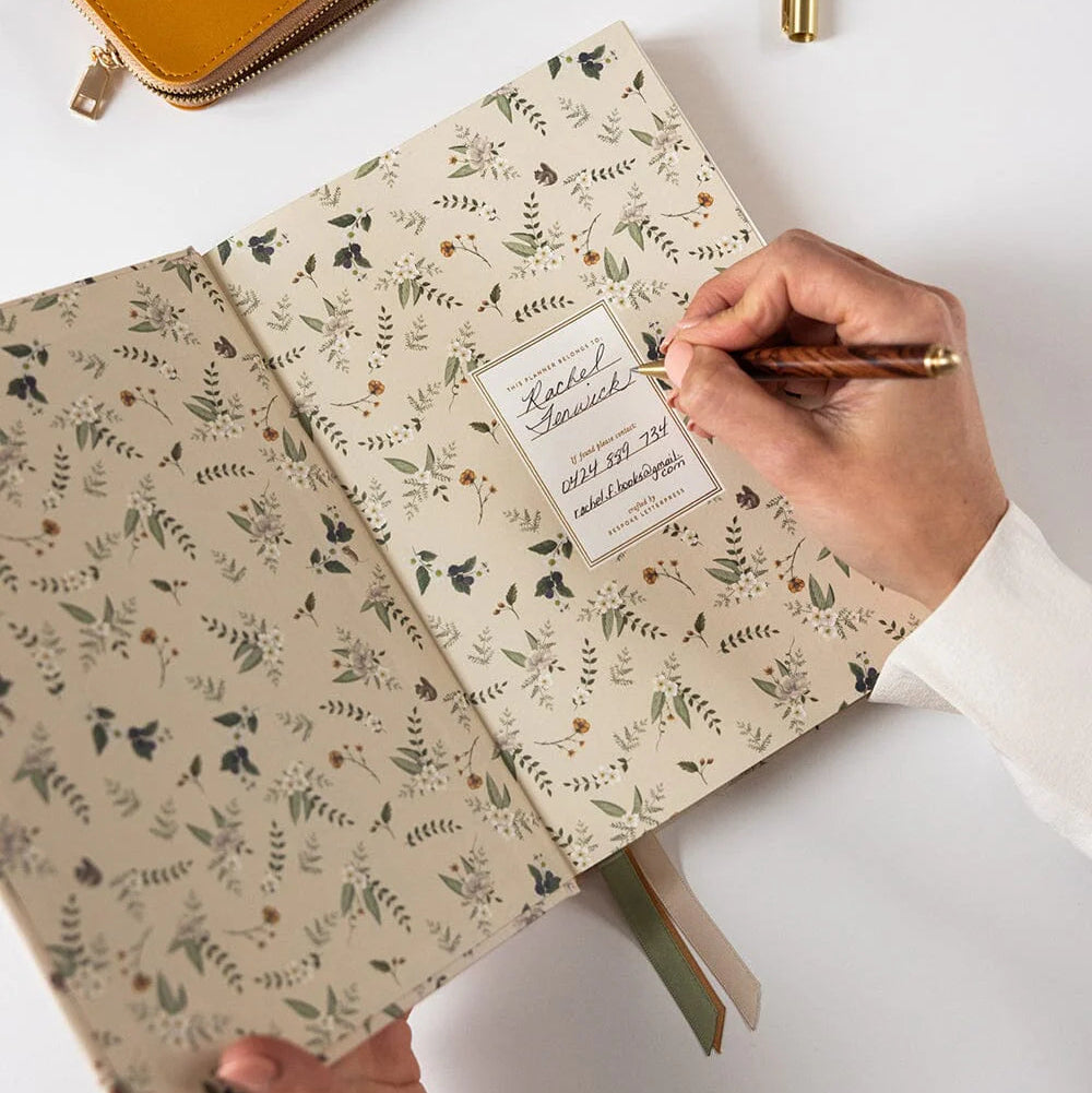 Person holding a floral notebook with a pen, on a white surface with a yellow pouch and pens.