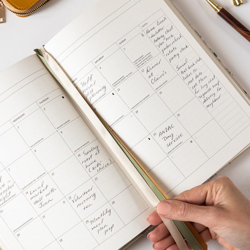Open planner with handwritten notes on a white surface, held by a hand with a pen.