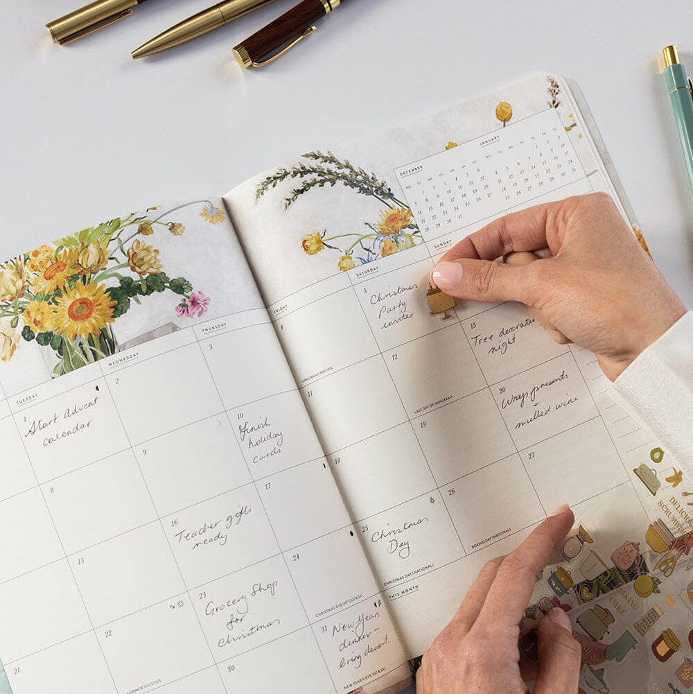 Person using a planner with floral designs and a hand holding a small object.