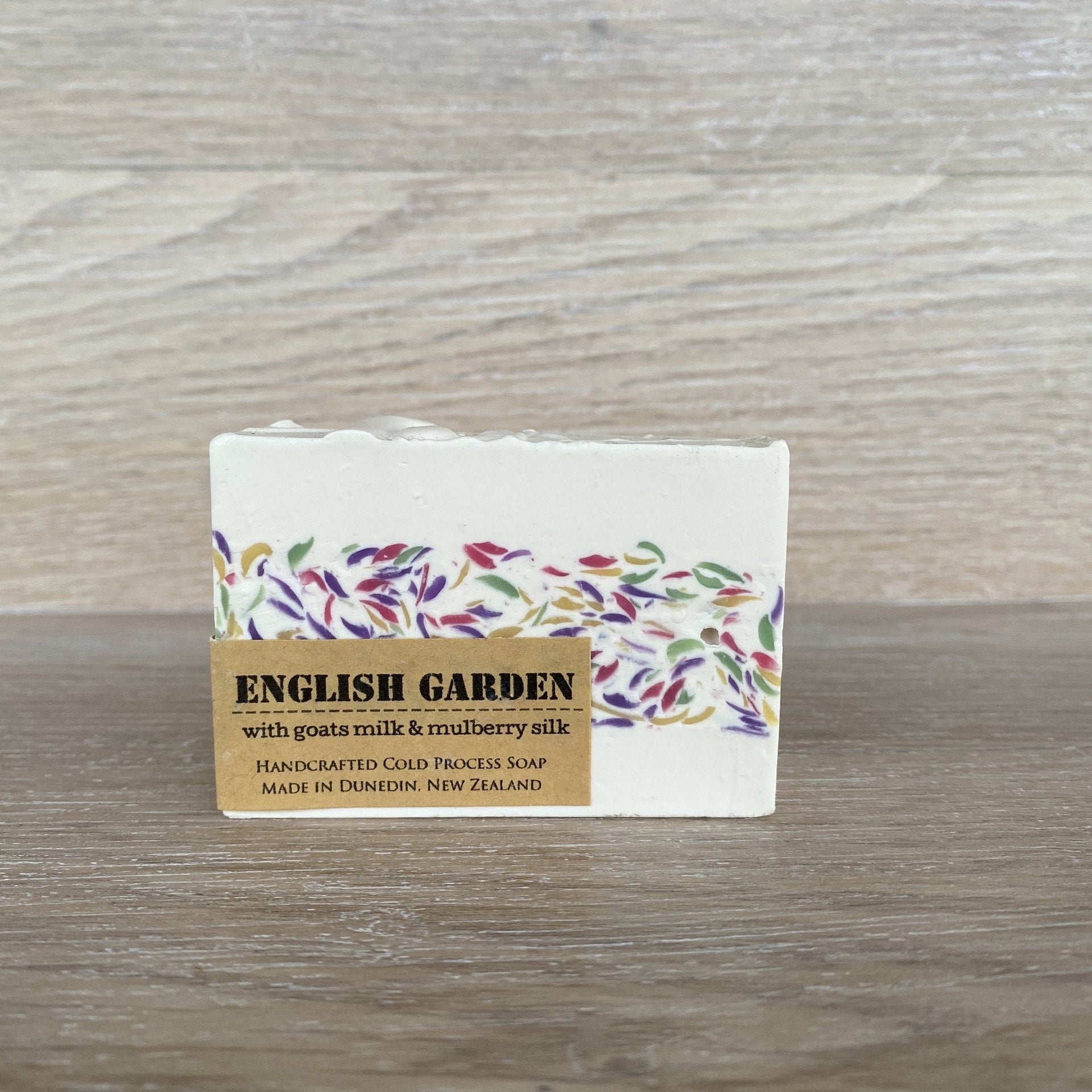 A rectangular bar of English Garden soap with a white base, a colorful floral pattern band, and a tan paper label, placed on a light wood surface.