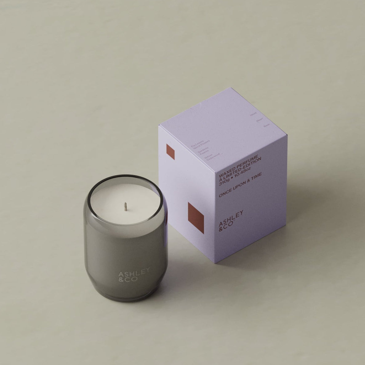 A dark gray glass jar candle next to its light purple packaging box on a neutral beige background. The brand name 'ABOLISH &amp; CO.
