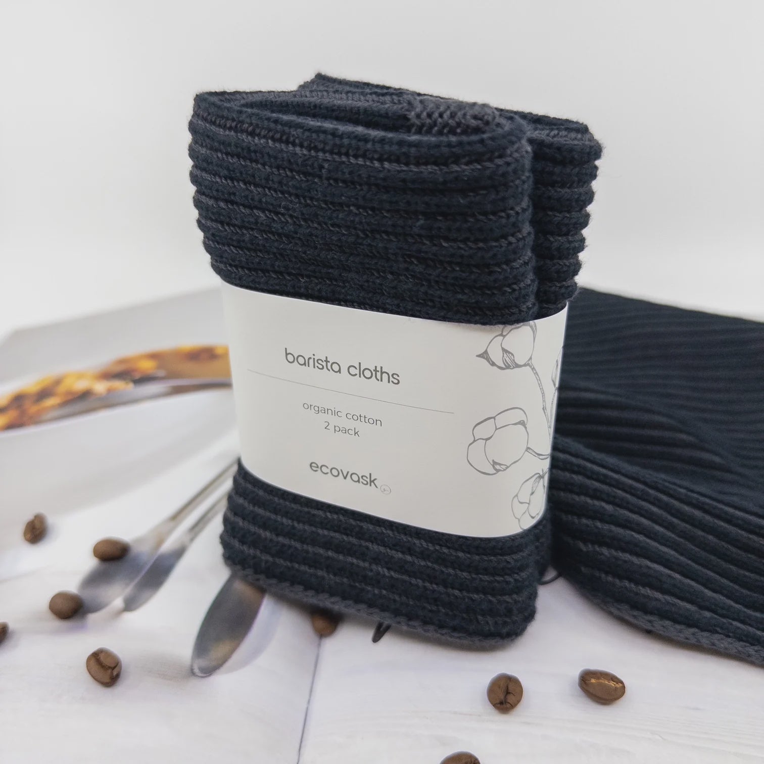 Pack of black barista cloths with a label on a white surface with coffee beans and a spoon.