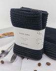 Pack of black barista cloths with a label on a white surface with coffee beans and a spoon.