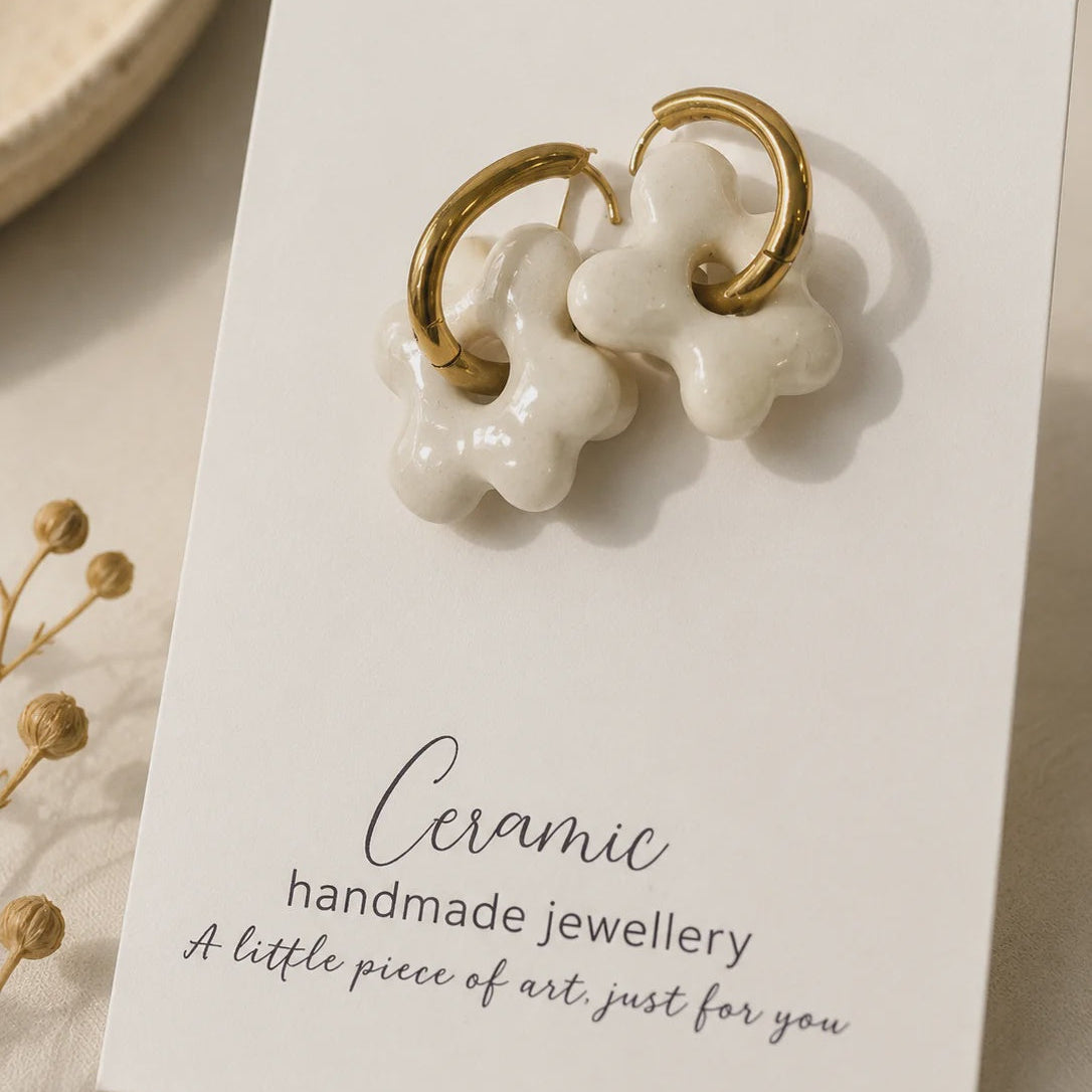 A pair of handmade ceramic stud earrings shaped like bone charms with gold-tone rings, displayed on a white card that reads 'Ceramic handmade jewellery'.