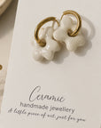 A pair of handmade ceramic stud earrings shaped like bone charms with gold-tone rings, displayed on a white card that reads 'Ceramic handmade jewellery'.
