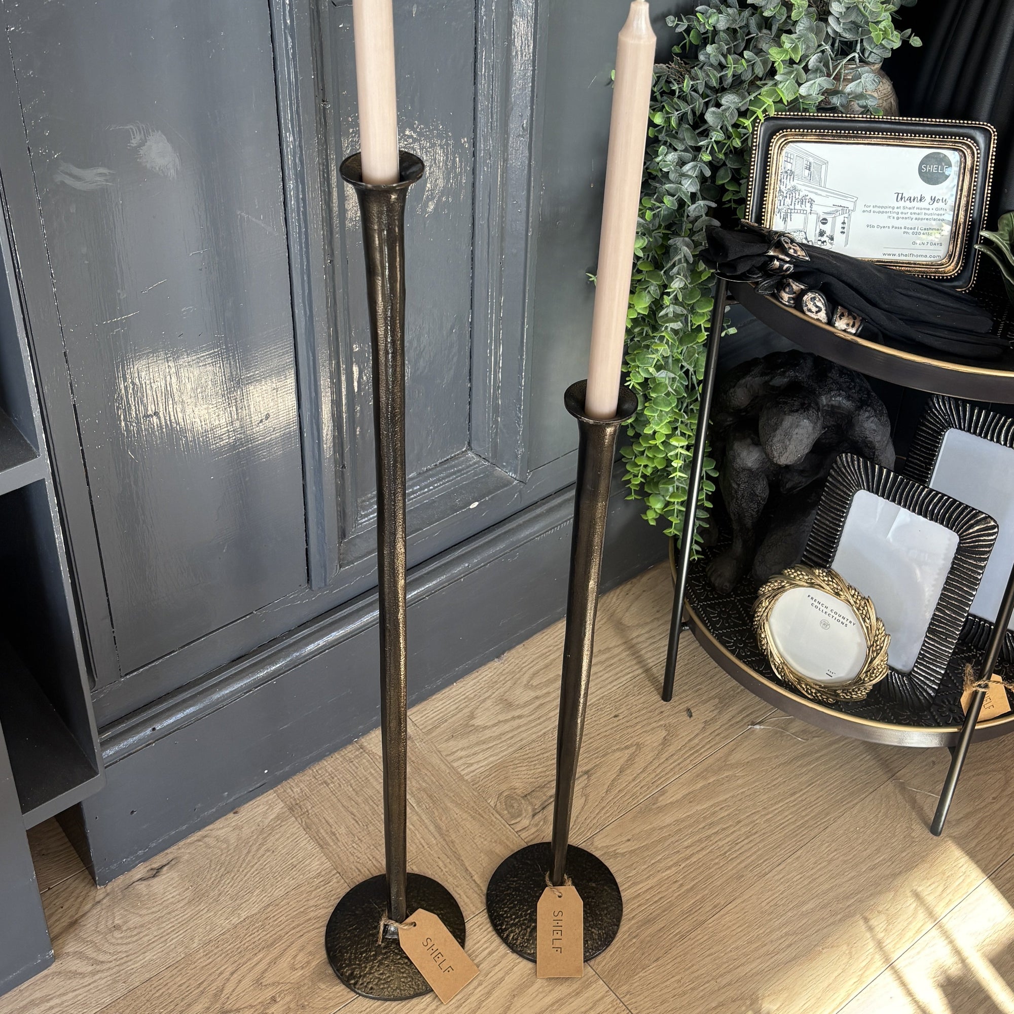 Two tall candlesticks with candles on a wooden floor in front of a dark gray wall.
