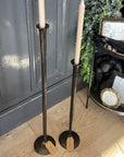Two tall candlesticks with candles on a wooden floor in front of a dark gray wall.
