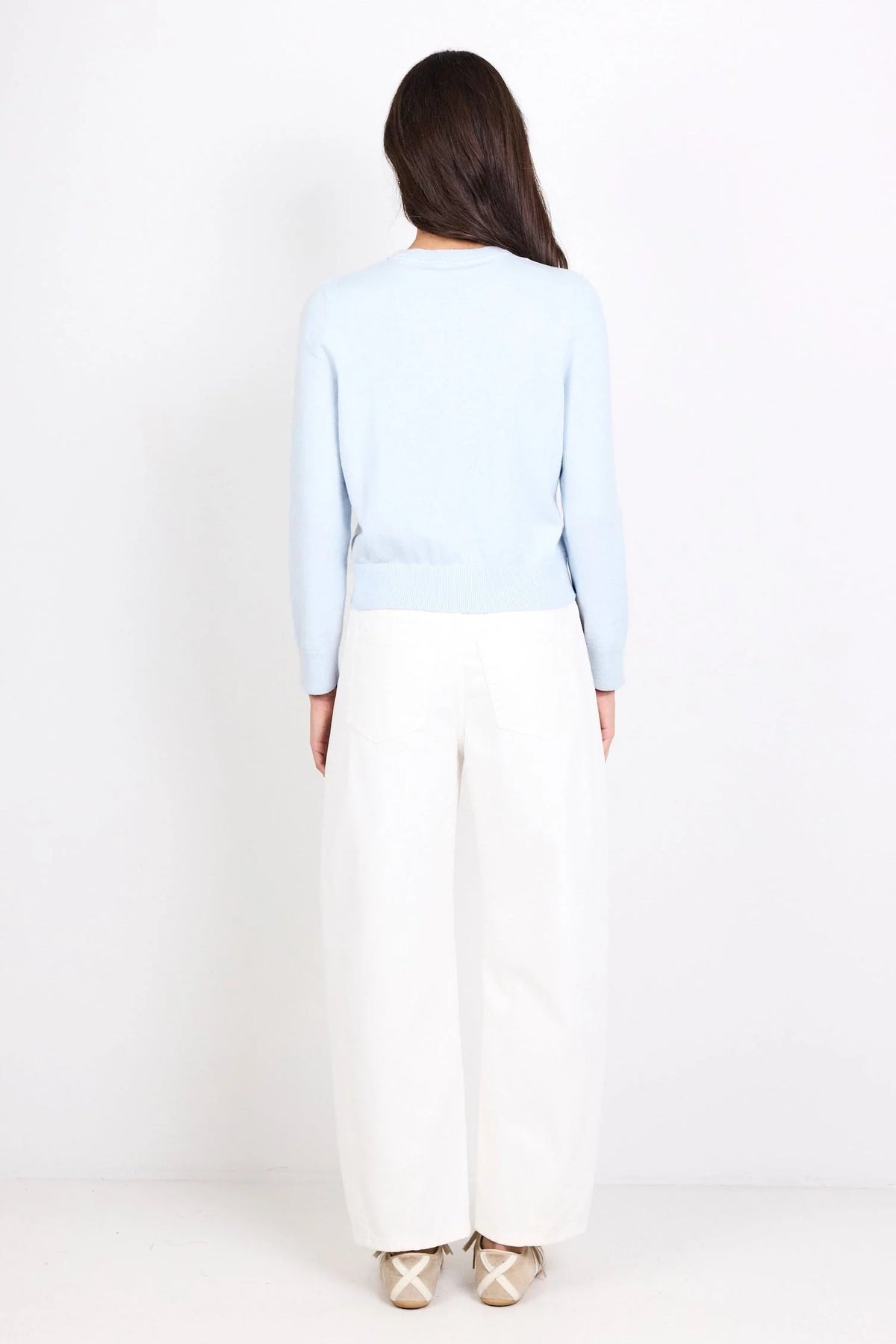 A woman shown from the back wearing a light blue long-sleeve knit top and white wide-leg trousers, standing against a plain white studio background.