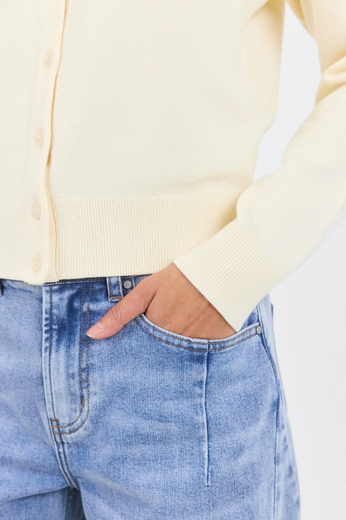 A close-up of a person wearing a pale cream cardigan sweater and light blue jeans, with the hand in the jeans pocket.