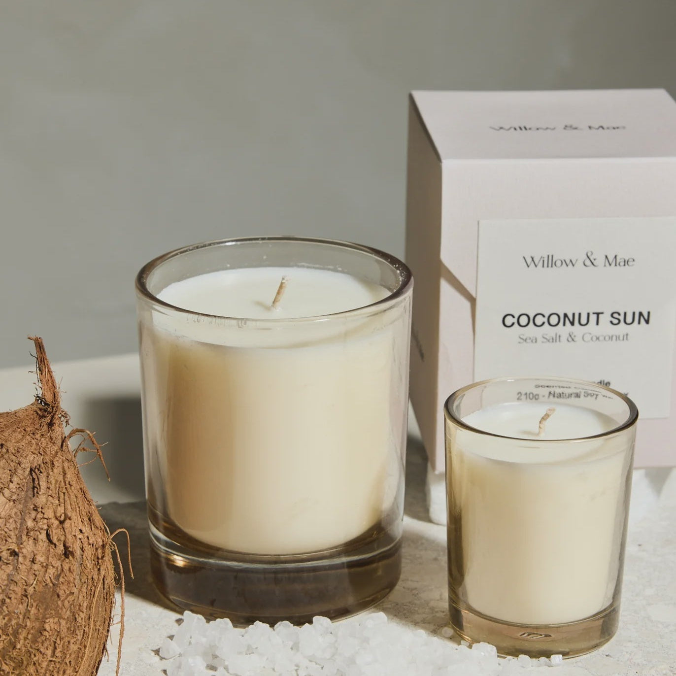 Two candles in glass holders with a box labeled 'Coconut Sun' on a textured surface.