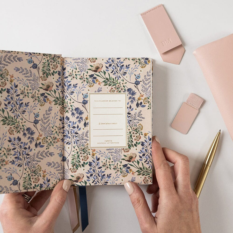 Floral-patterned notebook held by hands with a pen and pink case on a light background
