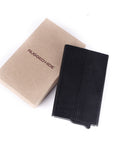 A black textured leather wallet or card holder shown next to its kraft paper packaging box, which is printed with the brand name 'RUGGED HIDE'.