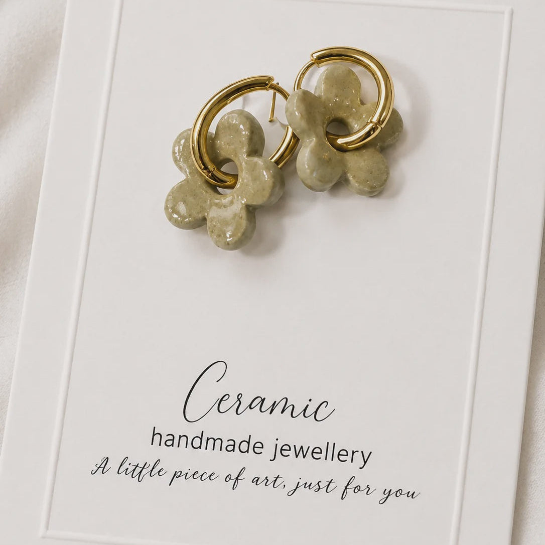 A pair of handmade gold hoop earrings with textured olive green ceramic flower charms, presented on a white display card.