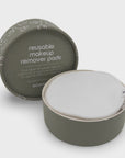 A reusable makeup remover pad in a round matte olive-green container, shown open with the white cotton pad inside. The matching lid sits behind it on a clean light gray background.