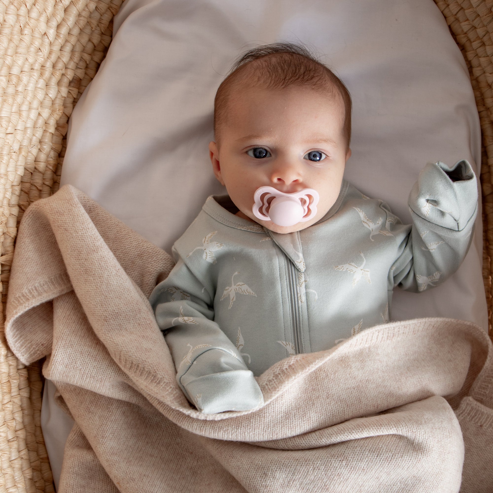 Baby in a light gray onesie with a pacifier, lying on a beige blanket in a woven basket.