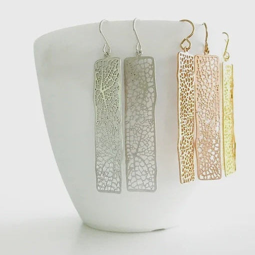 Three pairs of long rectangular dangling earrings with an open lace-like pattern, shown in silver, rose gold, and yellow gold. The earrings are displayed on a white ceramic cup against a clean white background.