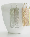 Three pairs of long rectangular dangling earrings with an open lace-like pattern, shown in silver, rose gold, and yellow gold. The earrings are displayed on a white ceramic cup against a clean white background.