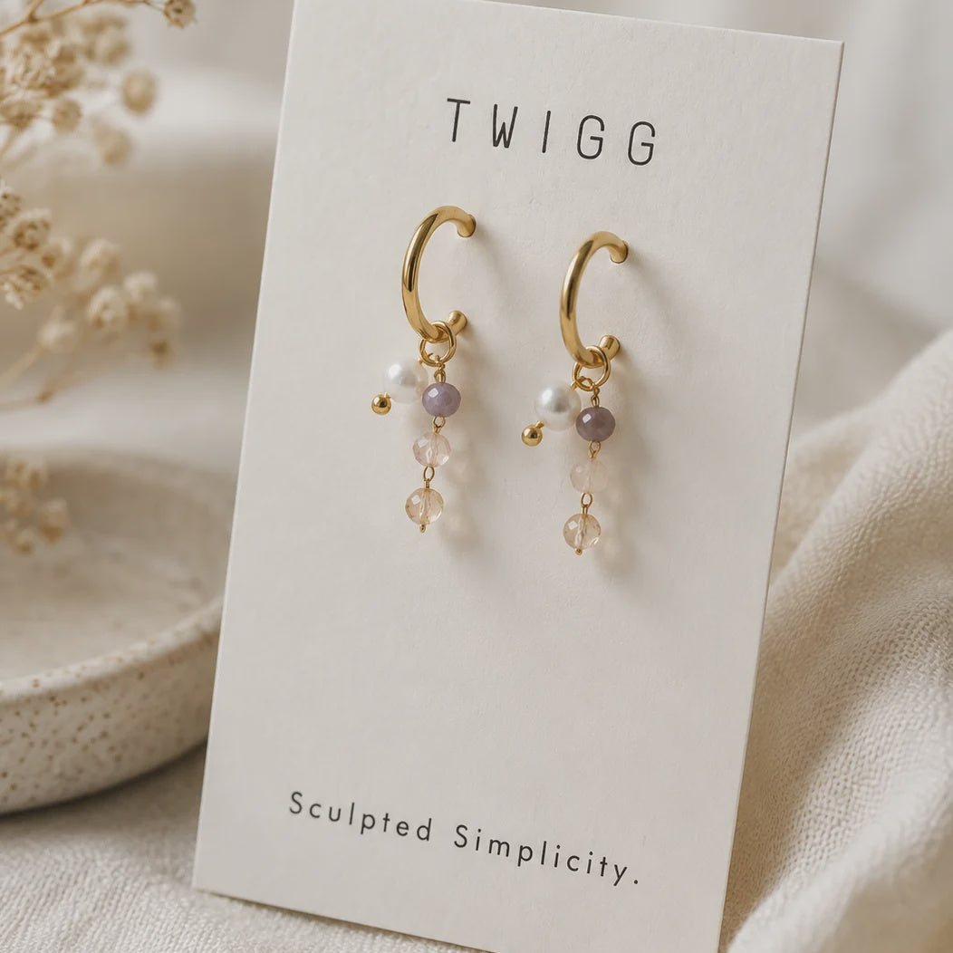 A pair of gold crescent hoop earrings with small pearl-like beads and pale lavender crystal drops, displayed on a white card labeled TWIGG, set against a soft cream fabric background with dried flowers.