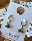 Gold and green earrings on a Foundry & Co card held by a hand.