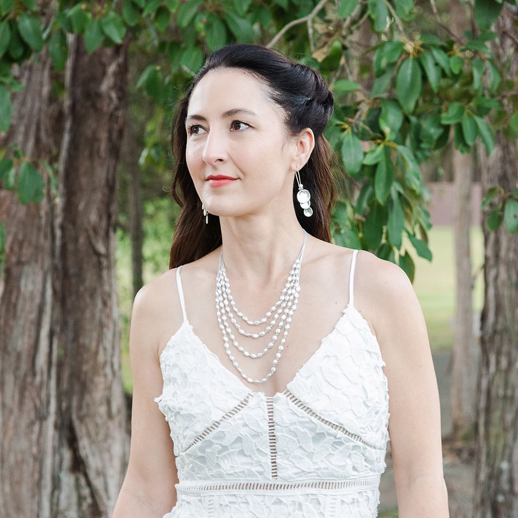 Woman in a white dress with pearl necklace and earrings standing in a natural setting.