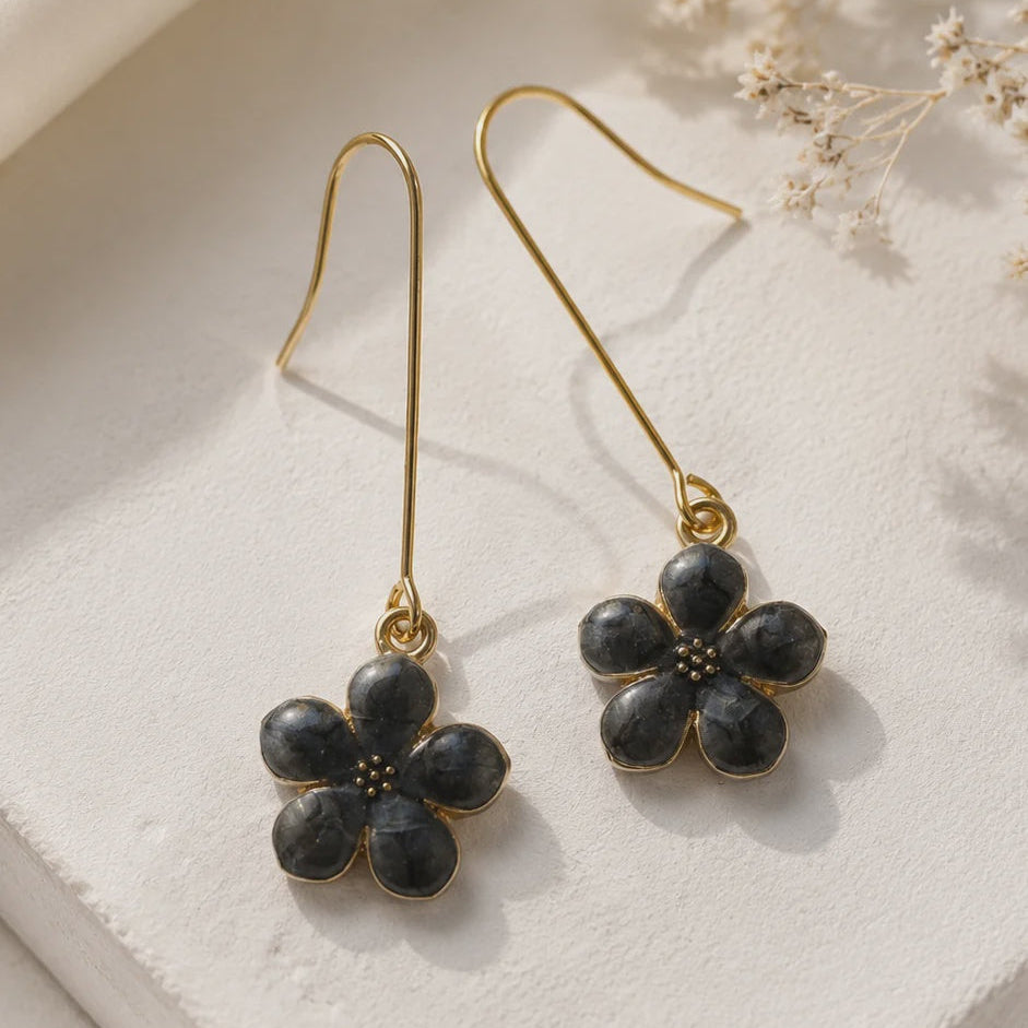A pair of gold-tone drop earrings with dark gray to black flower-shaped stone pendants, displayed on a textured white surface with dried flowers in the background.
