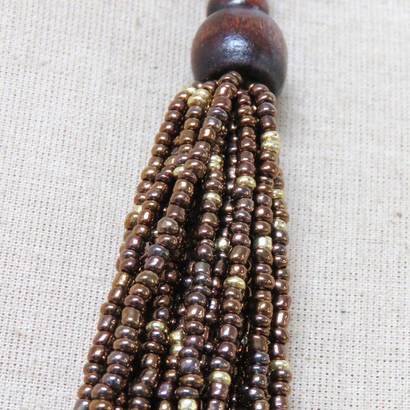 A close-up of a beaded tassel with a dark brown wooden bead top and multiple strands of metallic bronze and gold seed beads, laid on a beige textured fabric background.