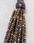 A close-up of a beaded tassel with a dark brown wooden bead top and multiple strands of metallic bronze and gold seed beads, laid on a beige textured fabric background.