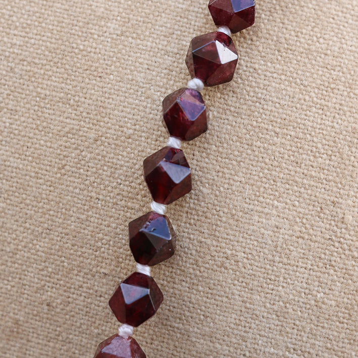 Faceted dark red gemstone beads strung on a white cord, displayed on a textured beige fabric background.