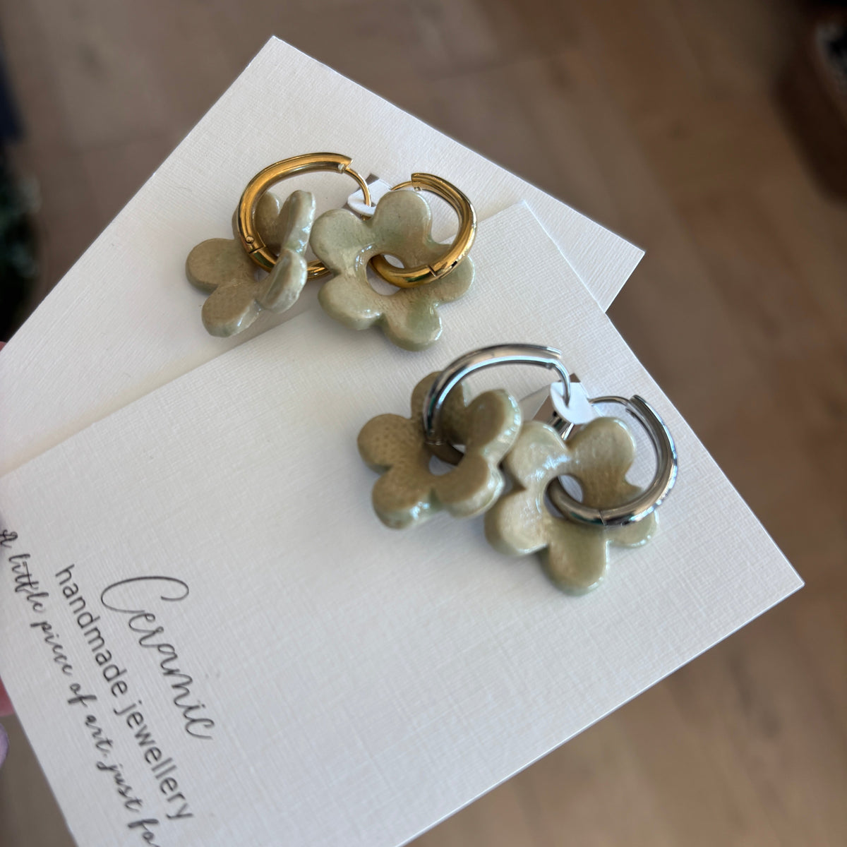 A pair of handmade dangle earrings displayed on branded white cards over a wooden floor. The earrings feature a light sage green clay flower charm paired with one gold hoop and one silver hoop.